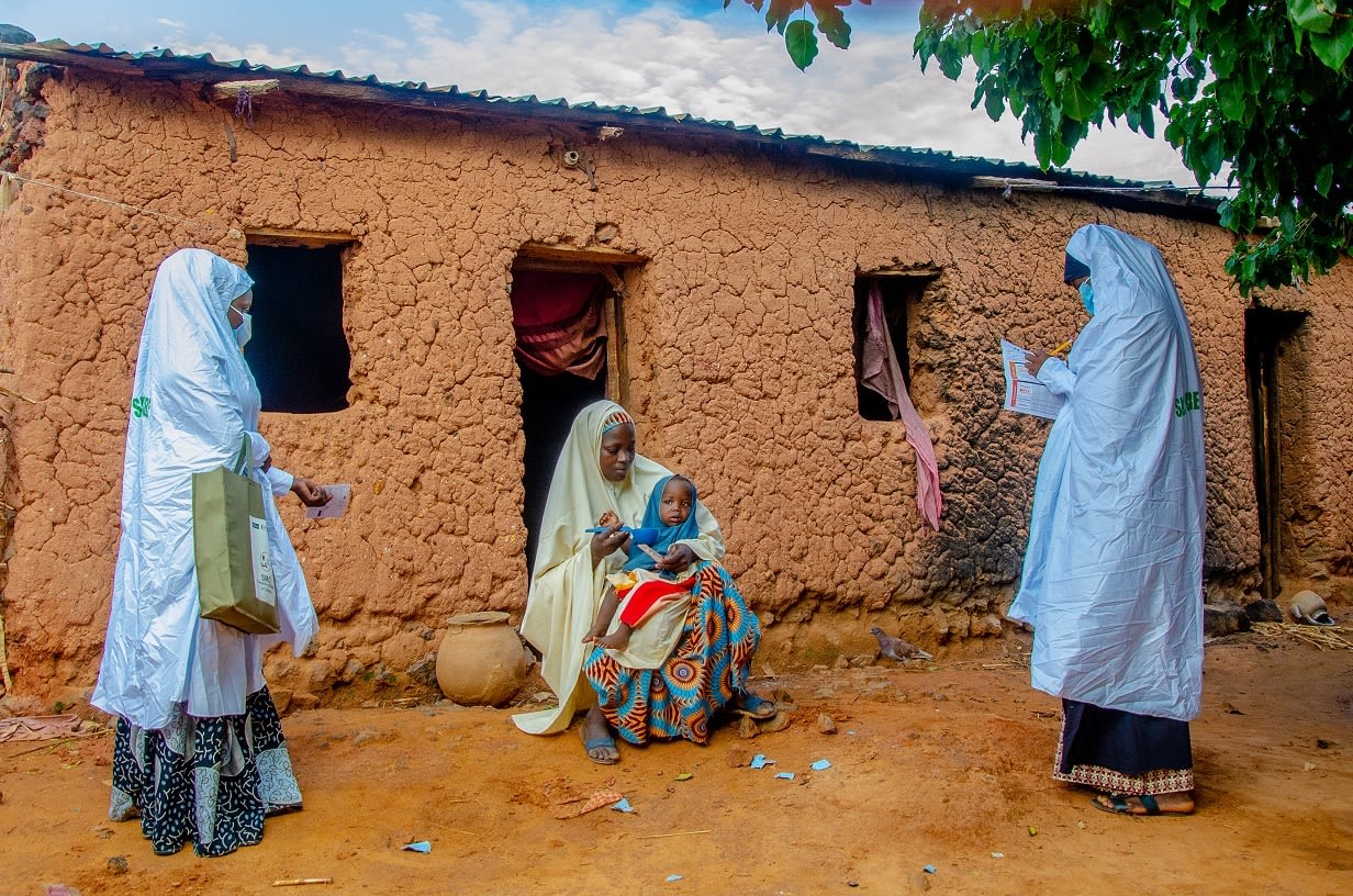 Two community health workers wearing PPE talk to woman seated outside her home holding a young child