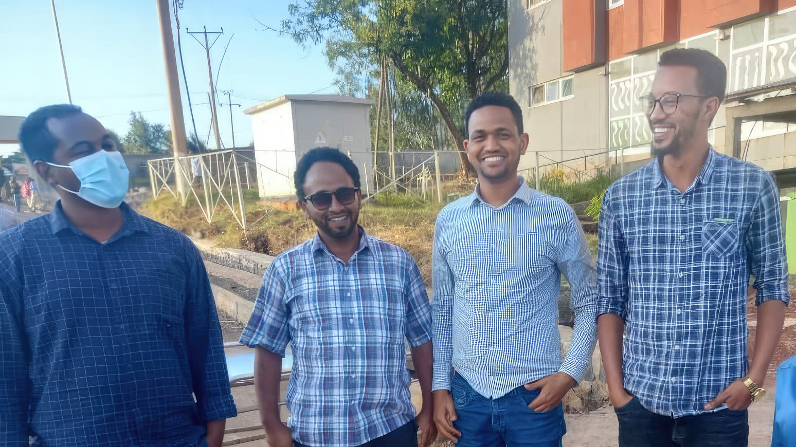 Four men smiling at camera, man on left wears surgical facemask