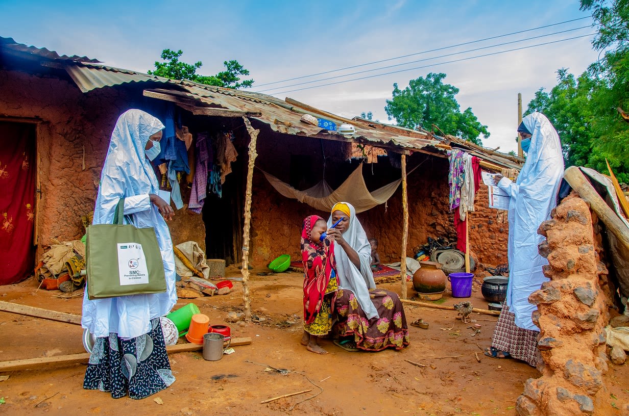 Two women wearing PPE stand watching seated mother holding cup to young child's mouth