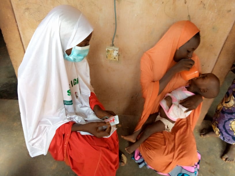 Health worker sits beside mother holding cup to mouth of small child