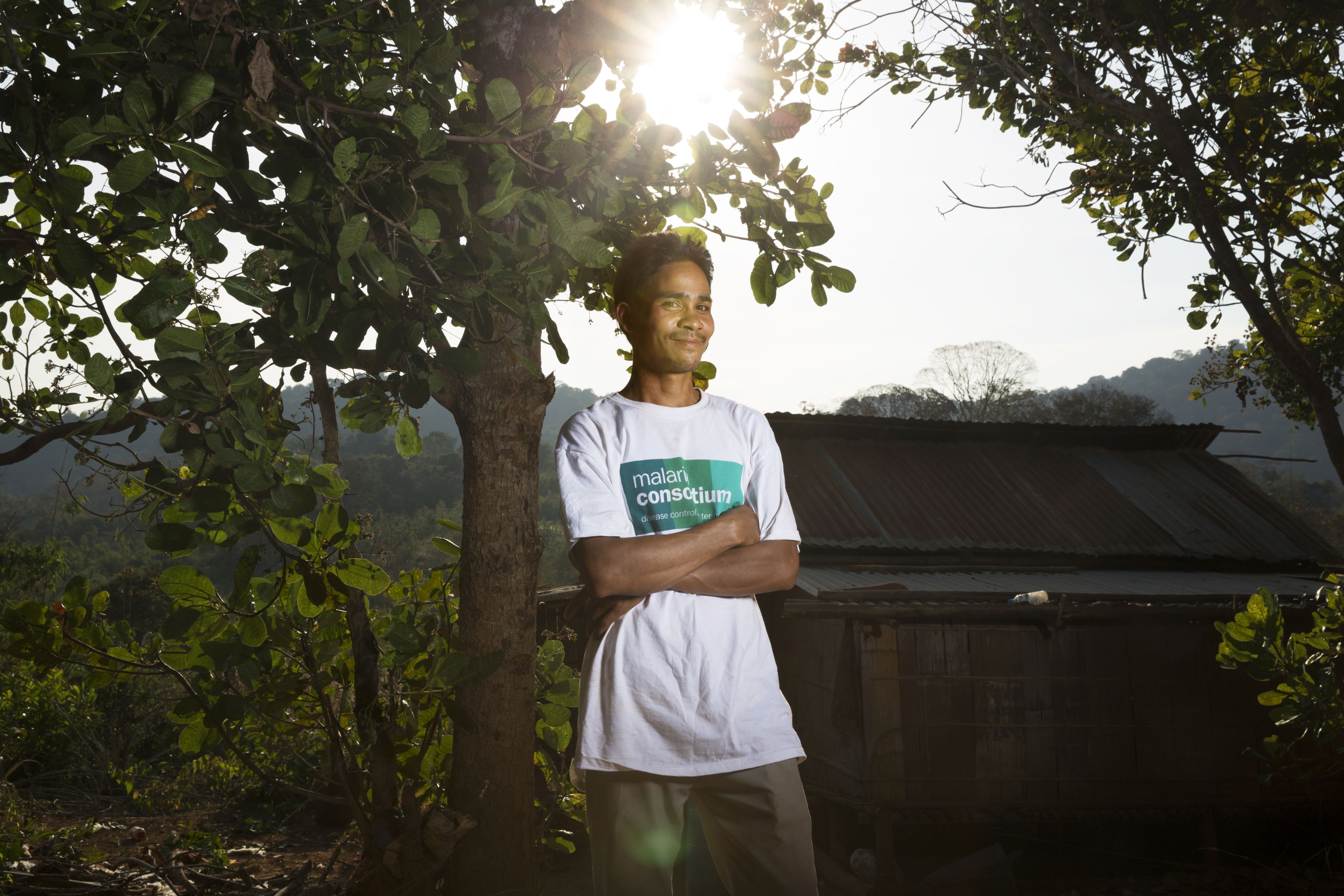 A young man wearing a Malaria Consortium t-shit stands in front of a home in a forest area