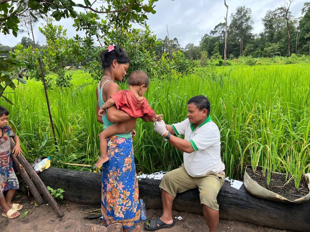 Mother holds young child as seated healthcare worker tests them for malaria
