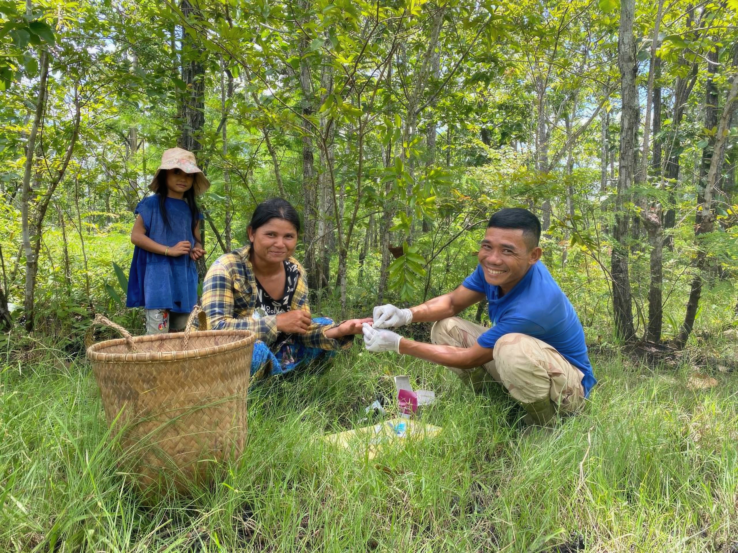 Smiling healthcare worker carries out malaria test on woman sat in grass as young girl stands behind
