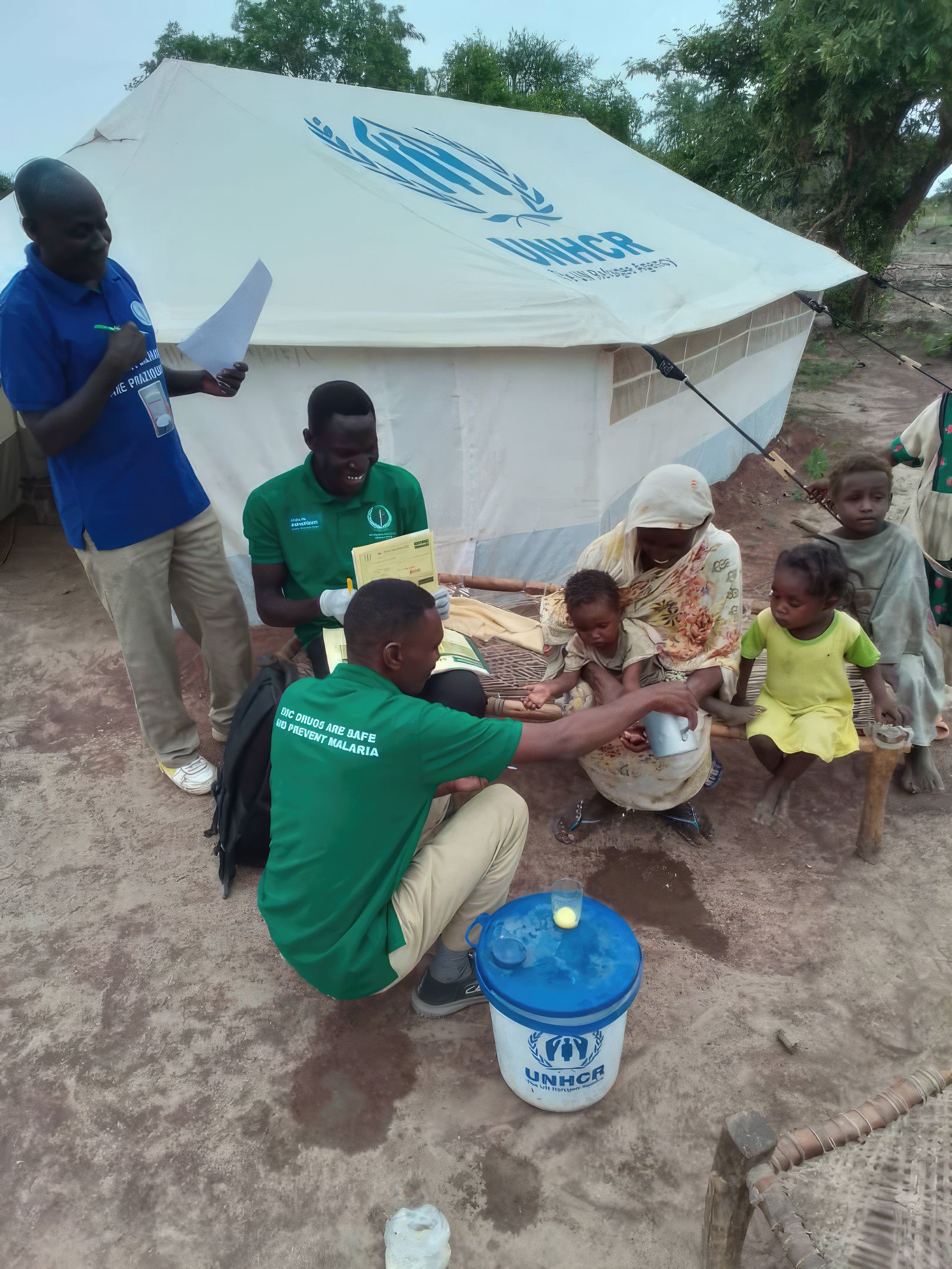 Smiling woman holds a young child as healthcare worker pours water from a jug to wash their hands