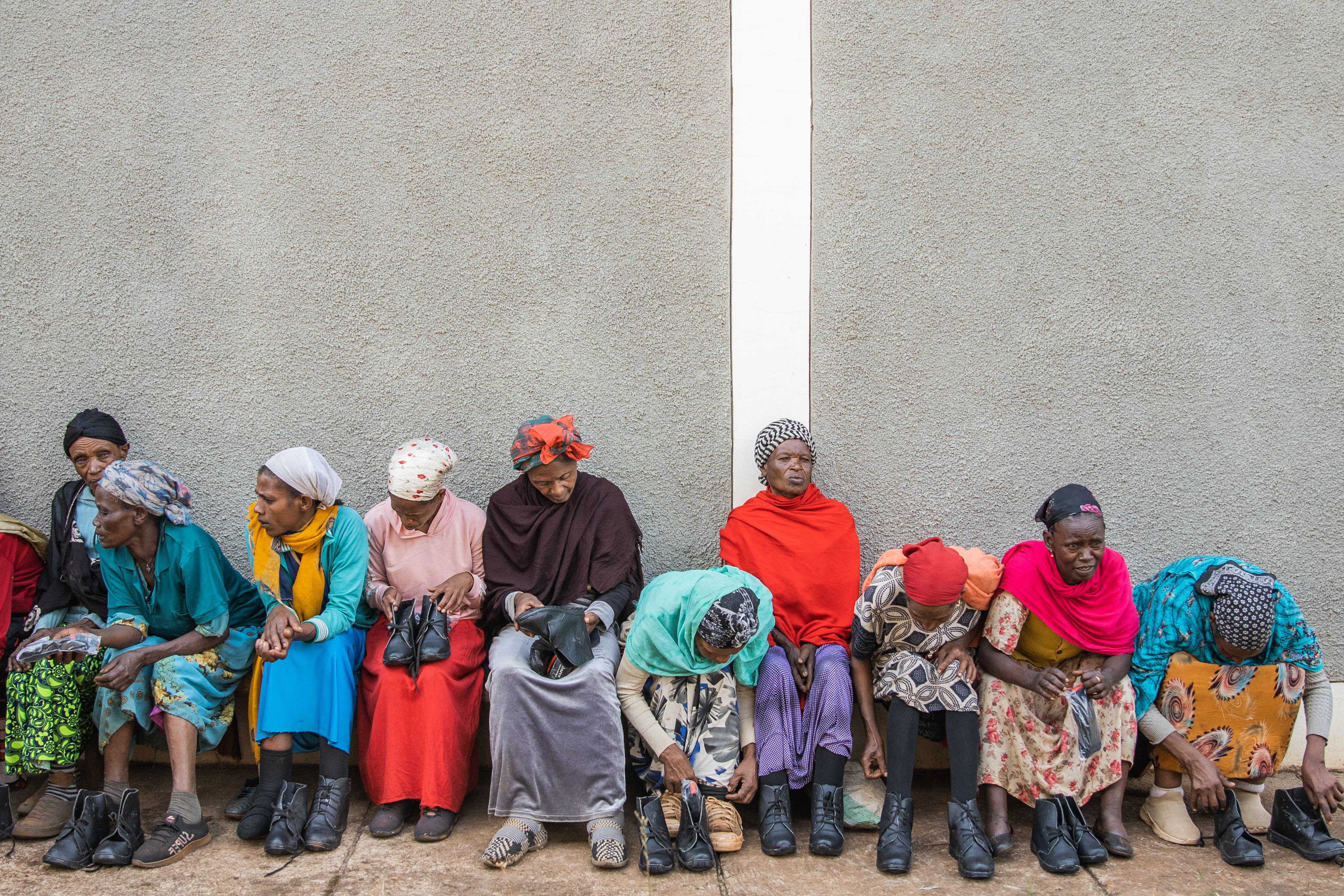 People sit on bench along wall with two women holding a pair of boots on their laps