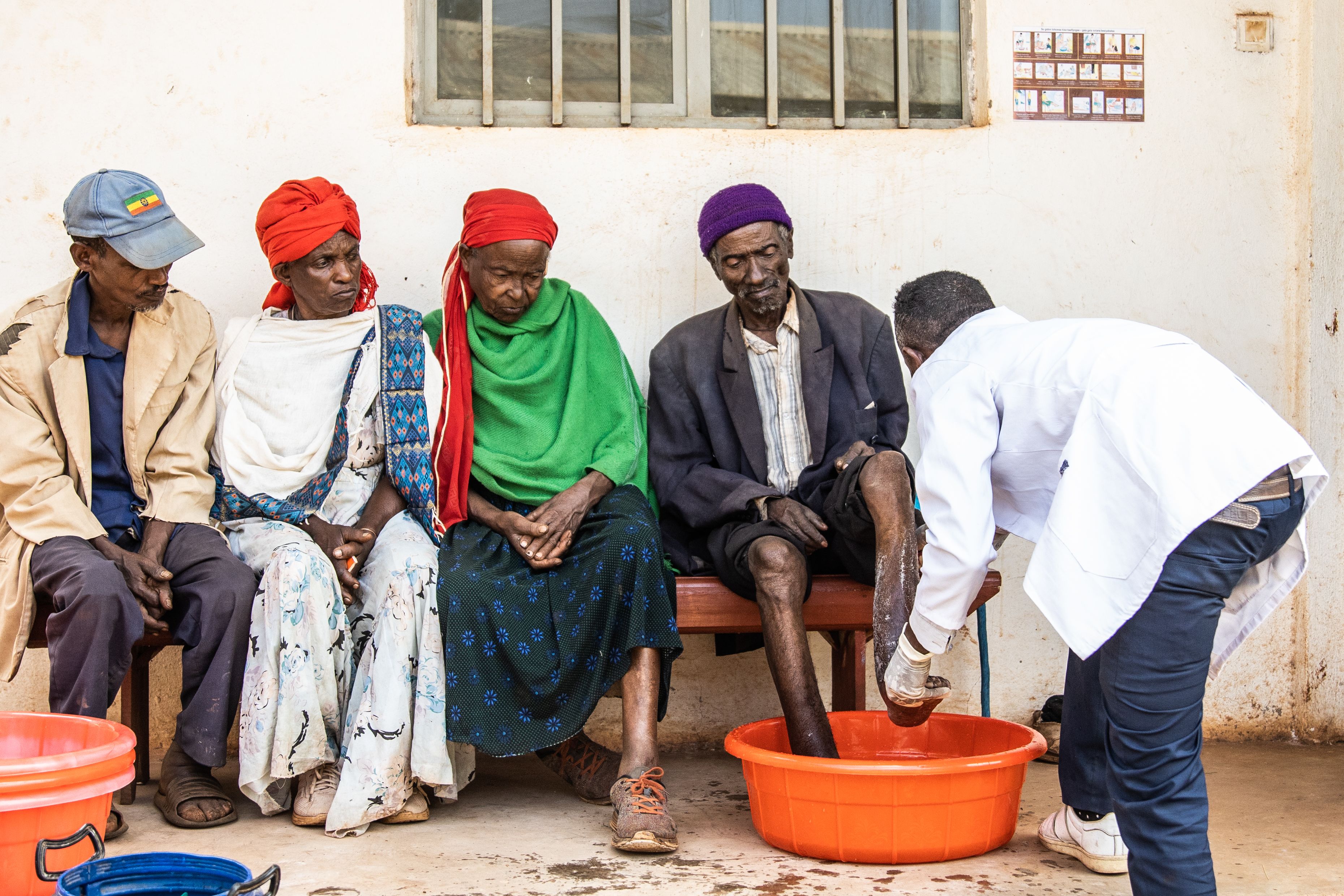 Healthcare worker washes man's feet in bowl as three people sit alongside watching