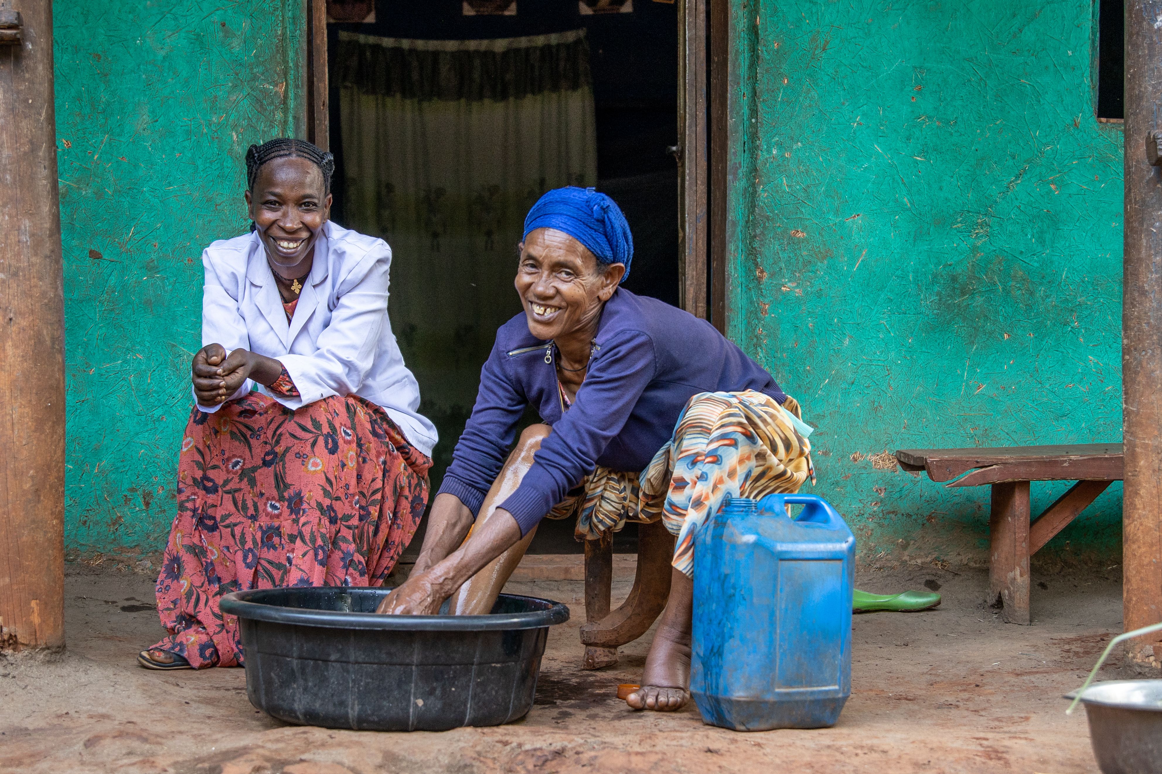 Two women sit smiling as one washes her feet in a bowl of water