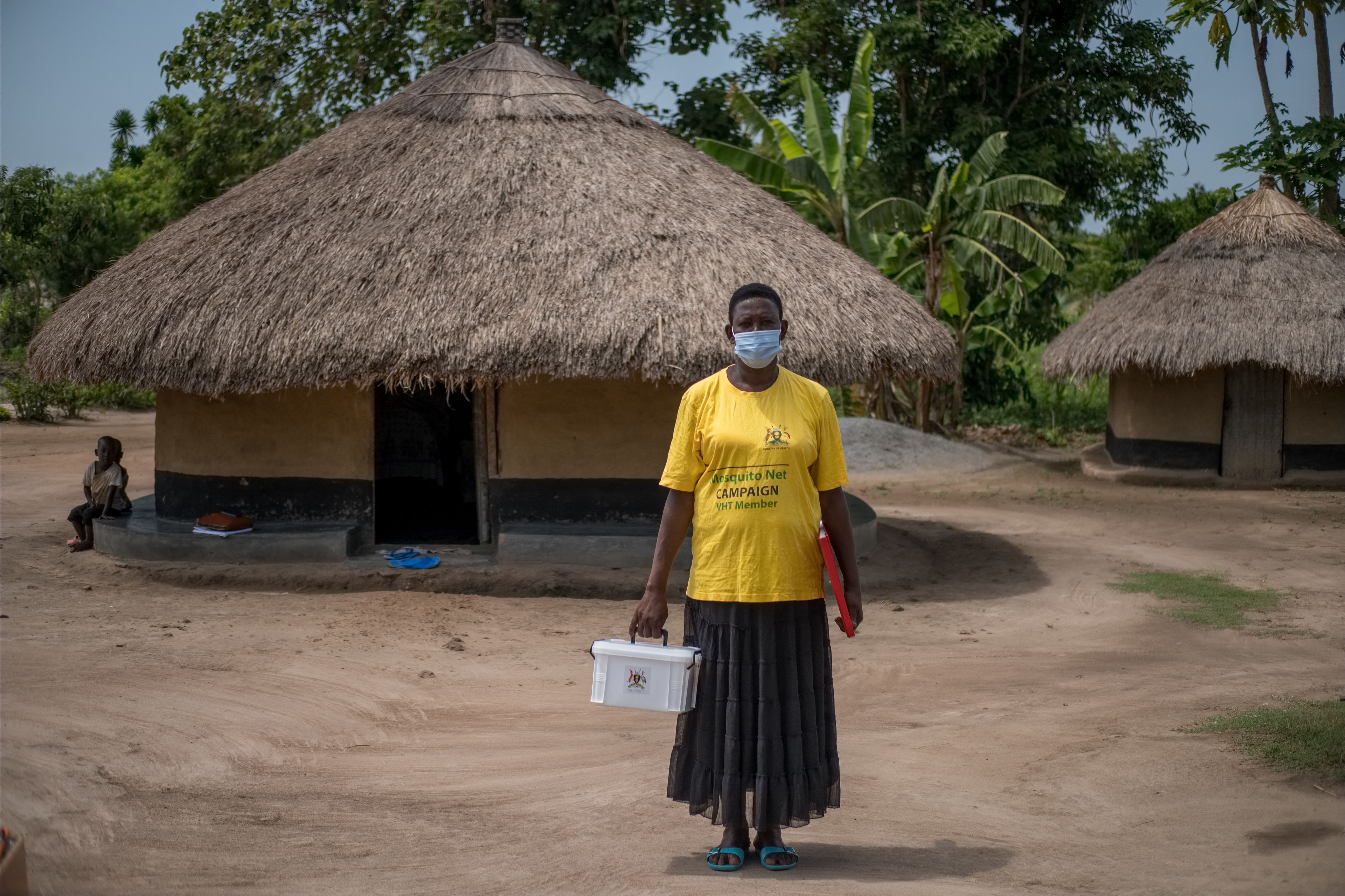 Health worker stands outside thatched hut holding a box of medicines