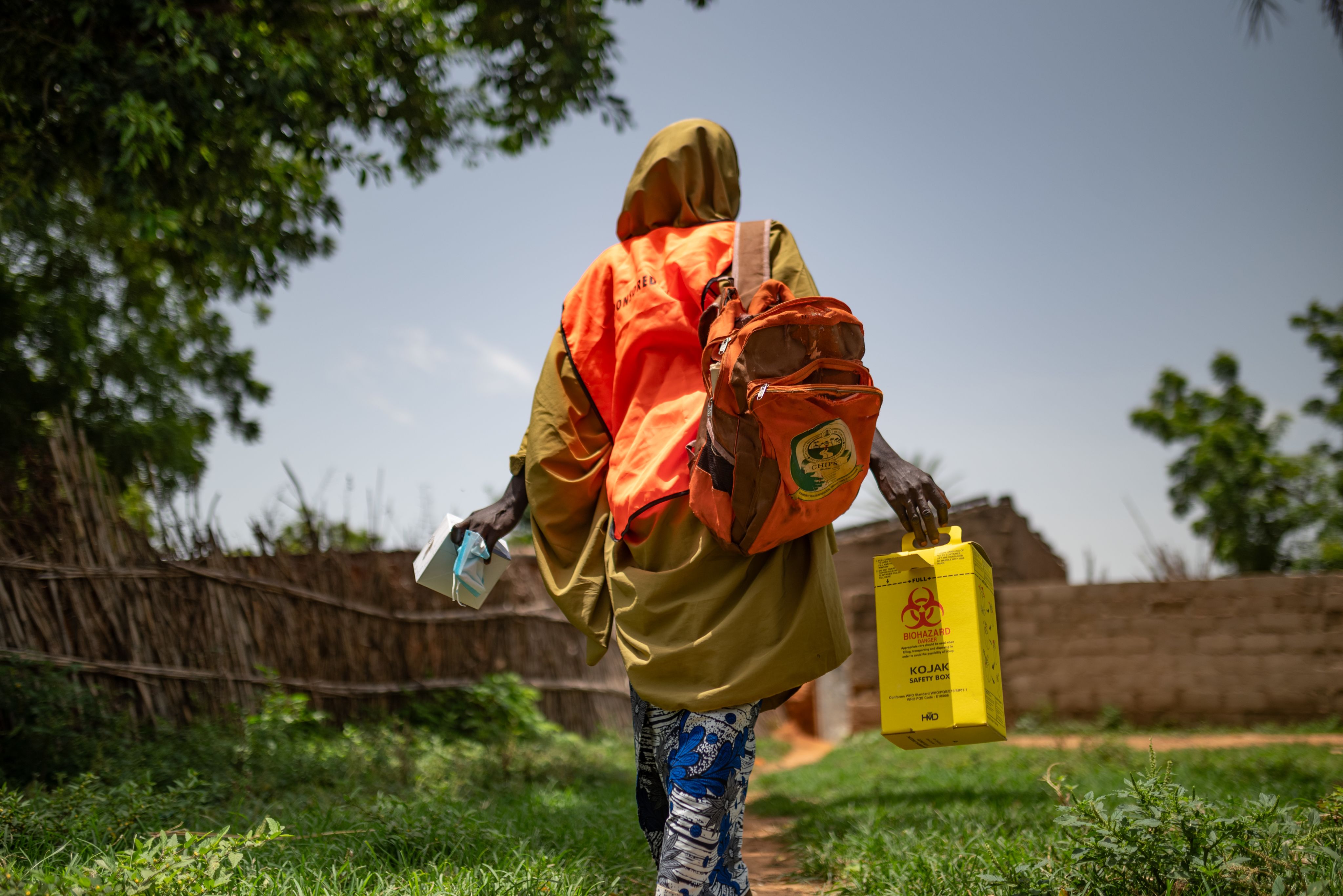 Woman in high-vis tabard with bag over her shoulder carries medical supplies towards house