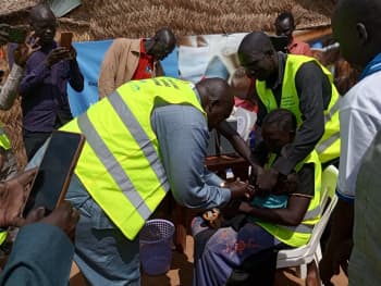 A small child sitting on someone's lap is given and injection by a man in a high-vis tabard surrounded by a group of people