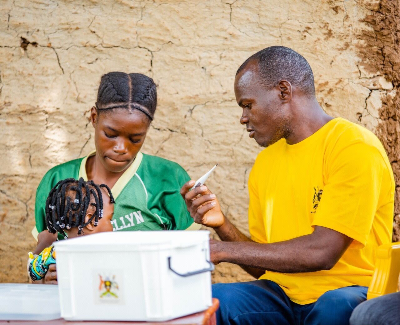 Man in yellow t-shirt holds thermometer as small child sits on mother's lap