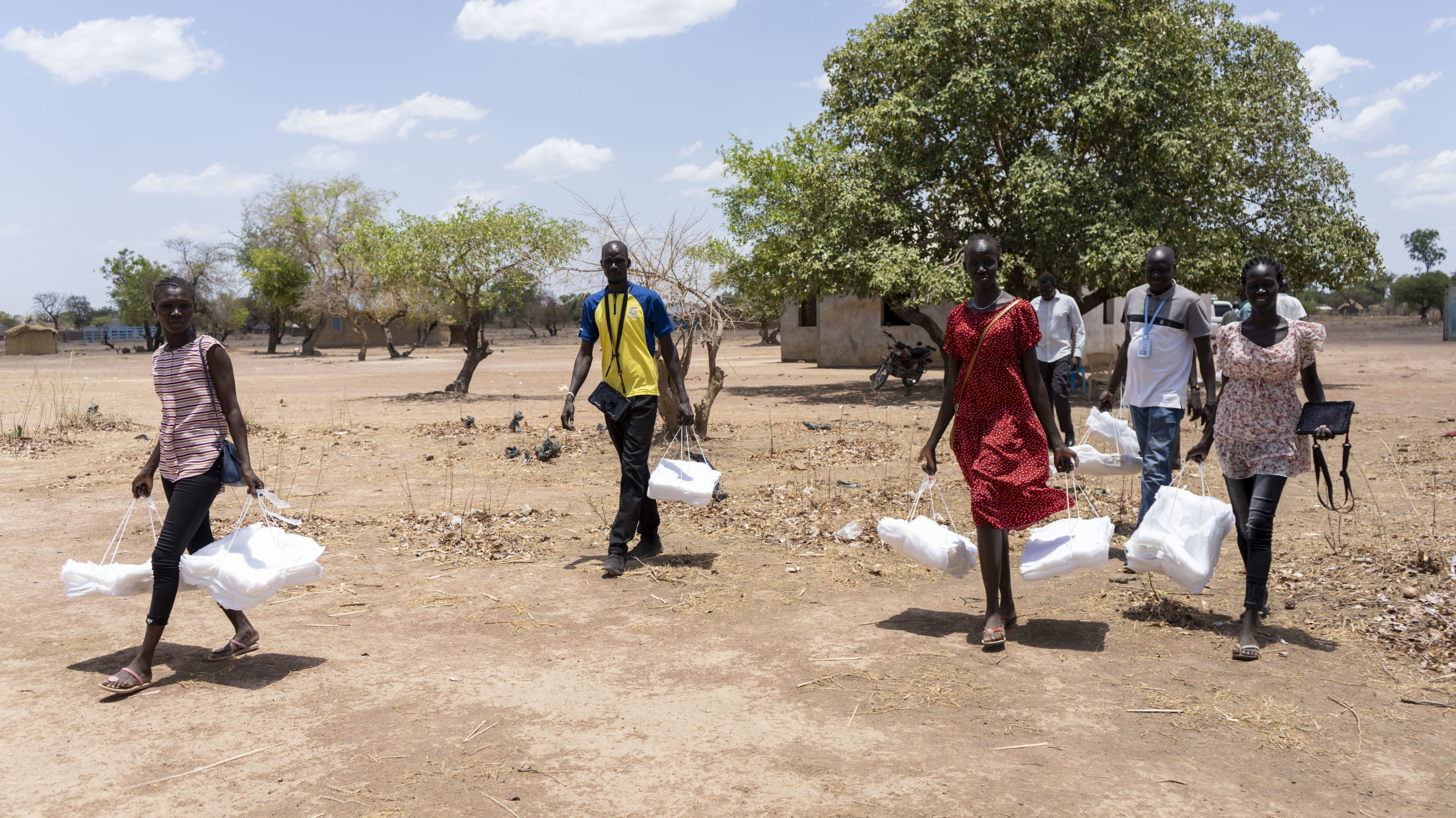 Group of people carrying bundles of mosquito nets walk across dry ground