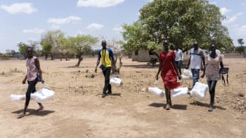 Group of people carrying bundles of mosquito nets walk across dry ground