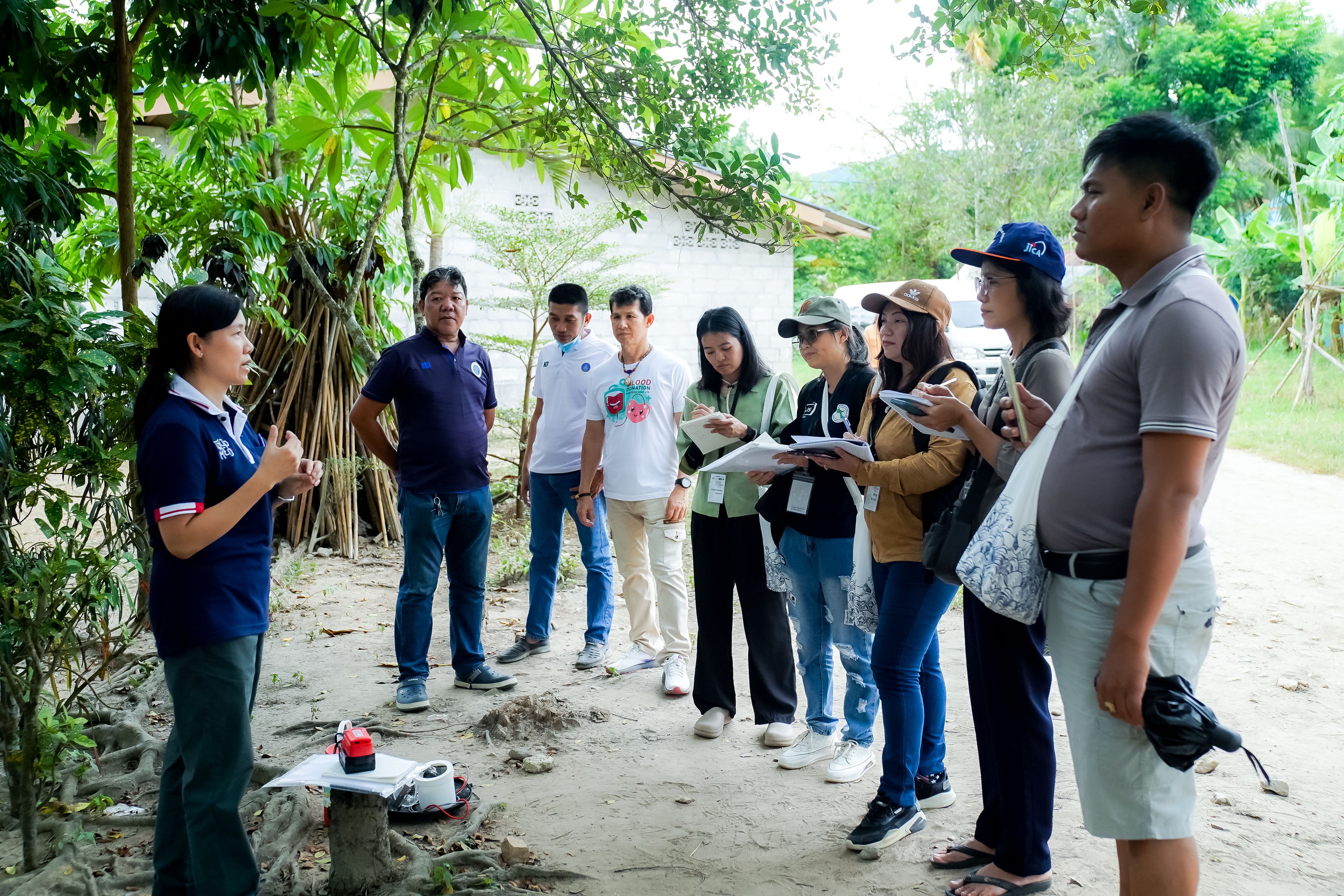 people stand outside under trees taking notes as they listen to a woman speaking