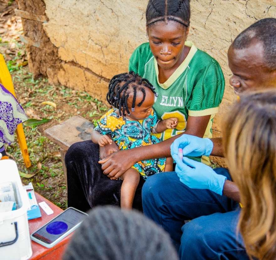 Man in yellow t-shirt wearing blue gloves prepares to carry out malaria test on small child sitting on mother's lap