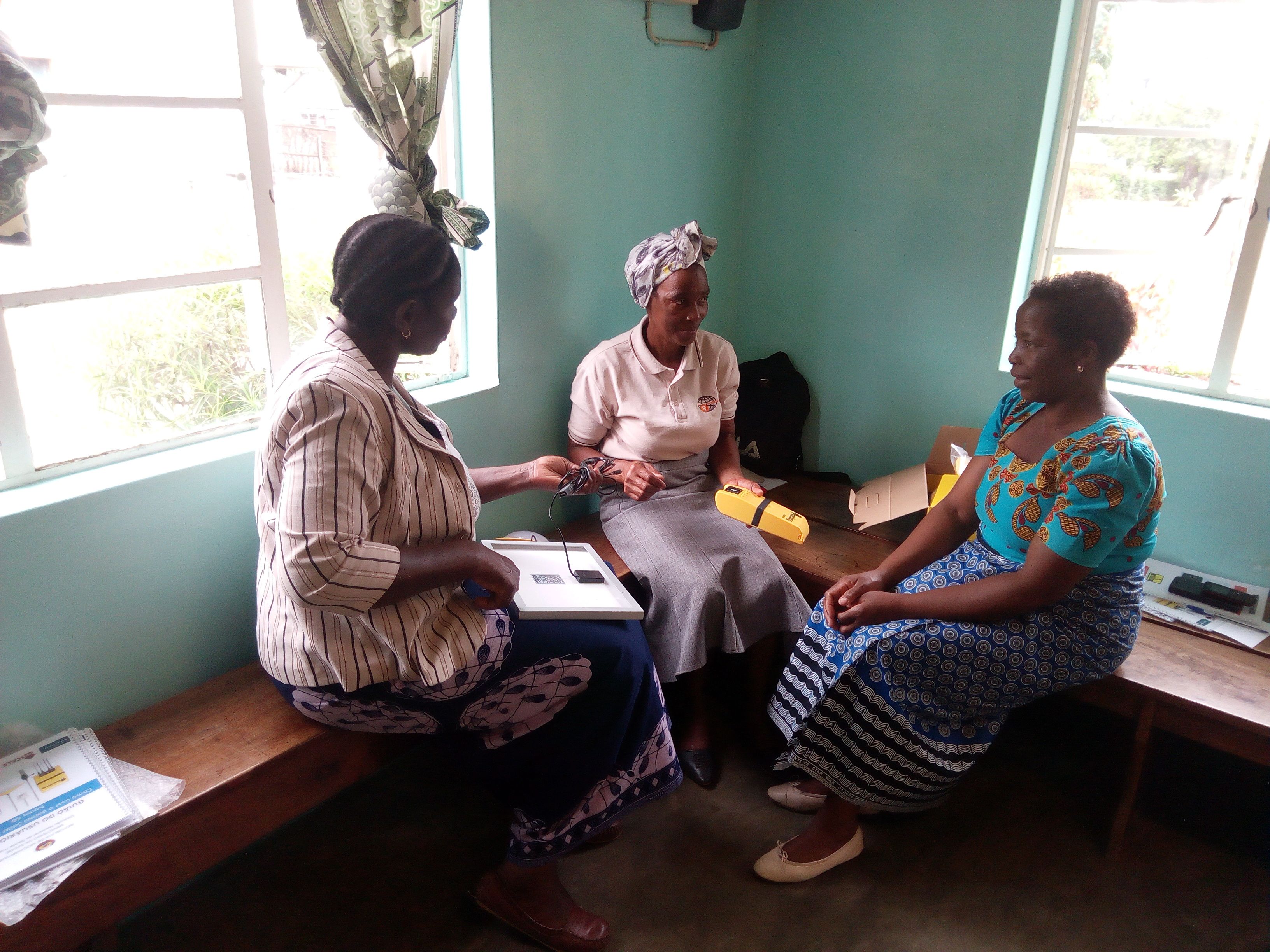 Three women sit on a bench in discussion; two hold solar panel testing equipment